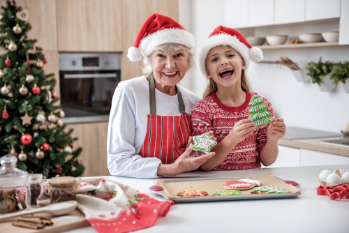 happy senior woman standing in kitchen with her granddaughter. They are holding baked colorful cookies and laughing