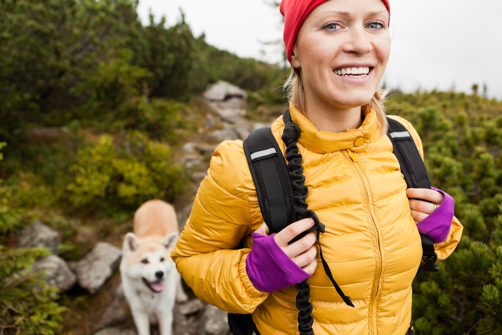 A woman in a yellow down jacket is hiking in the mountains smiling wide with a dog at her side
