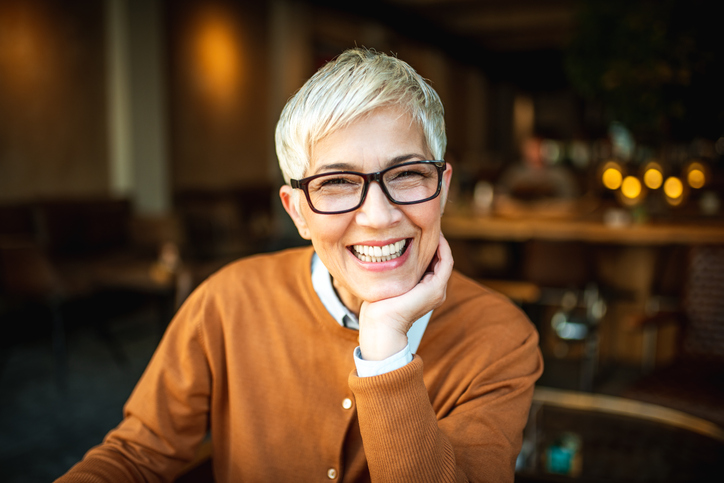 A middle aged woman with a blonde pixie cut black glasses and khaki fall jacket leans across the table with her chin on her hand and smiles at the camera