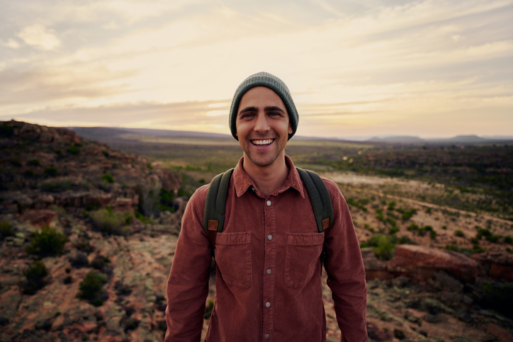 A young outdoorsy man in a gray beanie and red corduroy button down shirt smiles at the camera while standing in a desert landscape at sunsest