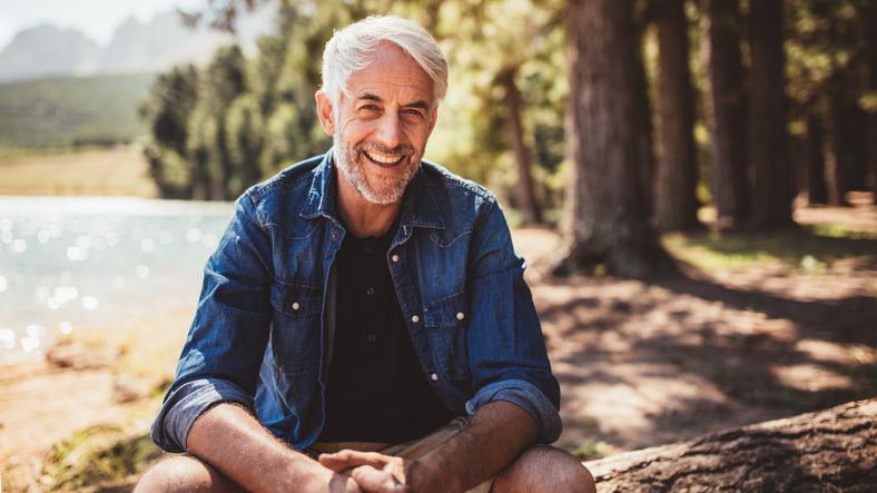 An older man smiling and sitting near a lake.