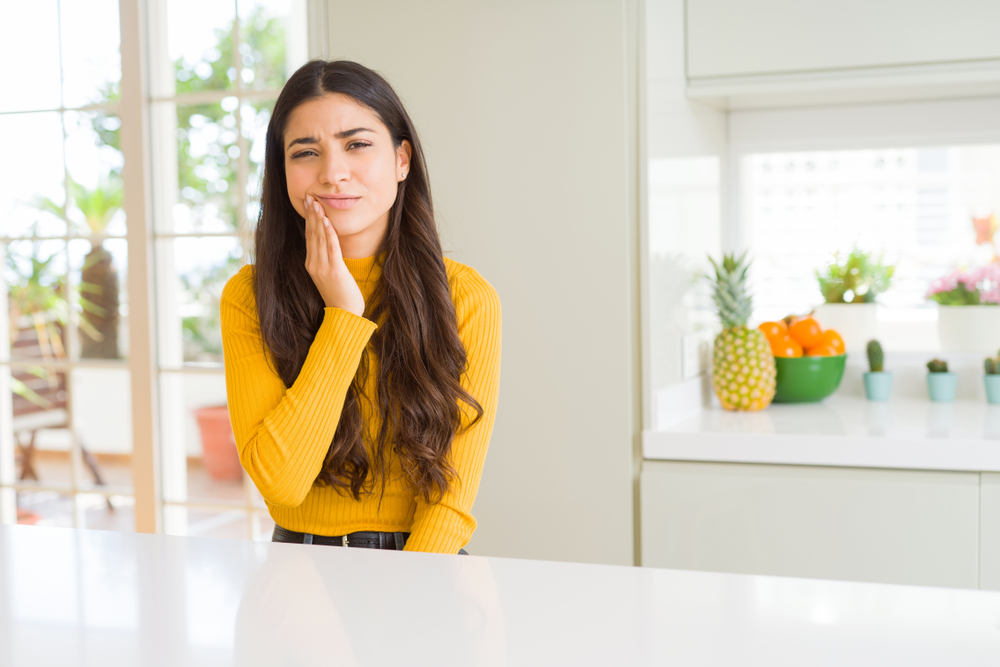 A young woman in a yellow top sits at the kitchen counter and holds her hand to her jaw in discomfort