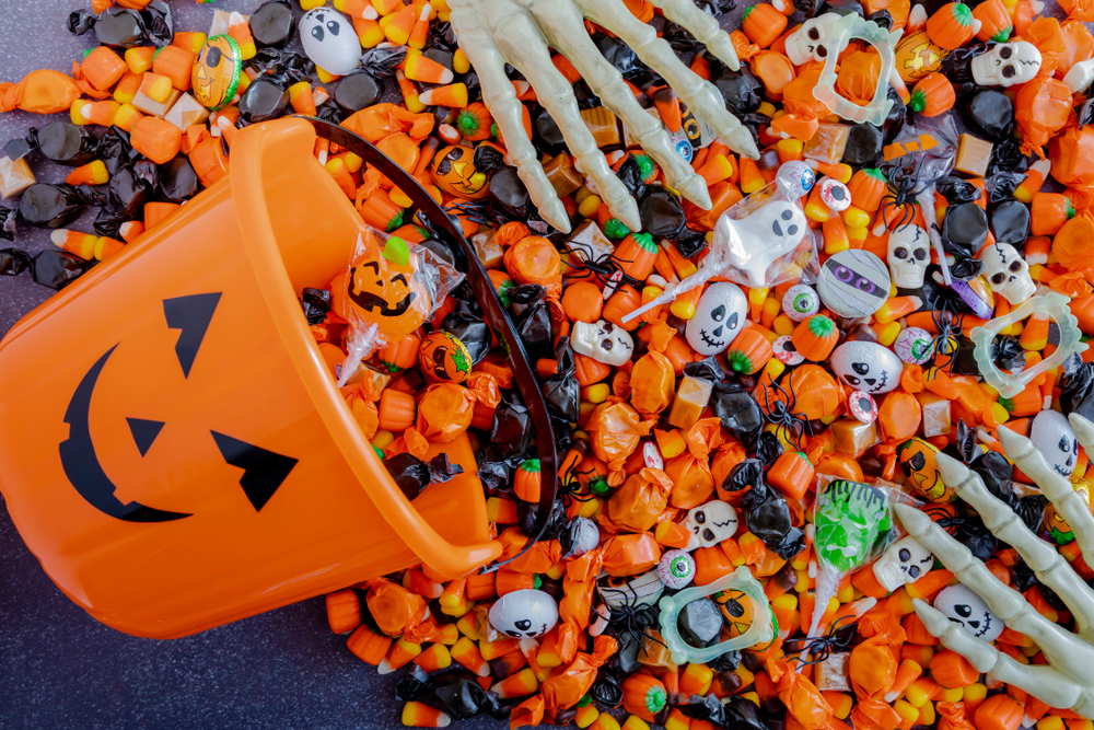 A close up of Halloween candies spilling out of an orange bucket with a pumpkin face and plastic skeleton hands grasp the candy